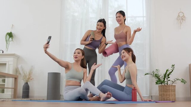 Smiling fit women in activewear taking photo with cell phone and greeting after exercise class at home, cheerful female friends sitting on yoga mats and making video call on mobile phone together