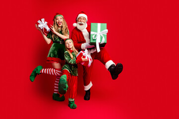 Naklejka premium Santa Claus and two cheerful elves presenting wrapped gifts and celebrating joyful Christmas family fun with laughing adults and child on a vibrant red background
