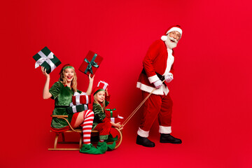 Naklejka premium Santa pulls a sleigh carrying two joyful elves with wrapped gifts in a festive family Christmas scene on a bright red background