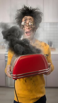Funny shock expression of young man with soot covered face and messy hair holding a smoking burnt red toaster in kitchen.