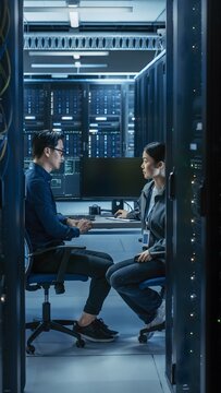 Two focused Asian IT system engineers sitting and discussing cloud network infrastructure inside high tech computer server room data center.