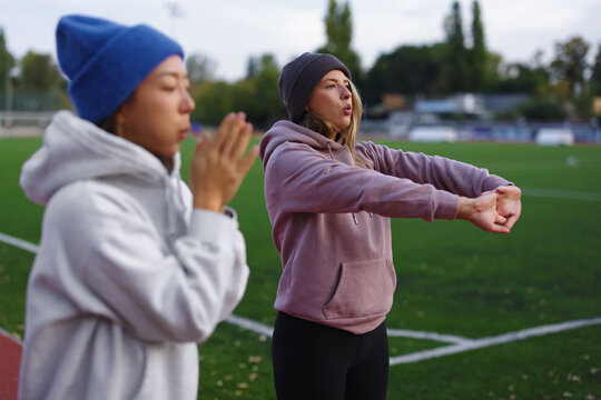 Two young diverse women warming up and stretching on an outdoor sports field, preparing for a workout, focusing on health, fitness, and active lifestyle