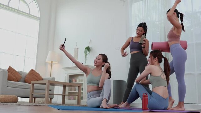 Group of happy young asian women in sportswear taking selfie with smartphone and waving hands after workout in studio, cheerful female friends sitting on yoga mats and making video call together