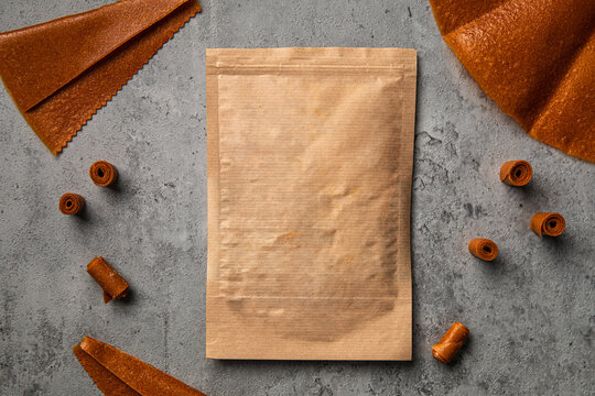 Natural fruit leather rolls and sheets arranged around craft paper ziplock pouch mock up on grey concrete background