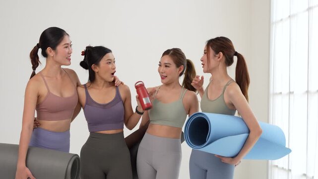 Group of happy young asian women in sportswear holding yoga mats and smiling after workout in fitness studio, cheerful young female friends talking and laughing together after exercise class indoors