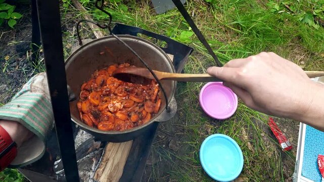 Man stirring frying carrots and onions in campfire cauldron for outdoor camping cooking lifestyle
