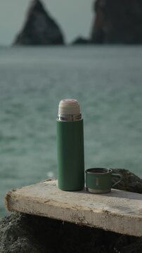 Thermos coffee coast green vacuum flask and cup rest on a weathered wooden board overlooking a tranquil ocean with distant rocky cliffs