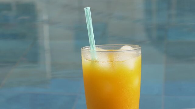 Chilled orange drink rests by swimming pool water, glass filled with ice and pale straw. Summer refreshment stands still in sunlit calm, suggesting leisure, cool hydration, and vacation ease.