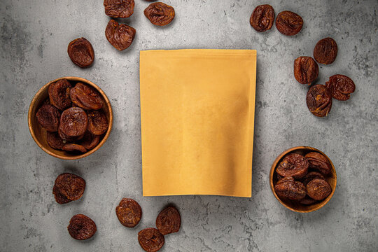 Natural dark dried apricots in wooden bowls and scattered around craft paper ziplock pouch mockup top view