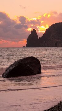 Dramatic ocean sunset crashing against a large coastal boulder at Fiolent beach, Crimea. Static medium shot.