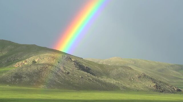 Rainbow arches above a green mountain ridge after rain over a bright summer meadow scene.