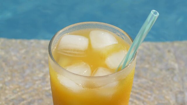 Close up of chilled orange beverage with ice cubes in clear glass and straw beside pool. Bright citrus drink looks refreshing and appealing, ideal for menu design, food photography, summer advertising
