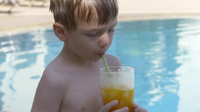 Boy drinks orange juice by pool, clutching tall glass with striped straw. Young child stays focused on sipping while standing in bright sunshine beside rippling blue water, holding beverage upright.