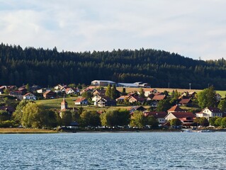 A picturesque European village with red-roof houses nestled along a serene lake shore, backed by a lush pine forest and rolling hills under a soft daylight sky, perfect for travel and rural themes. © MadameSayCheese