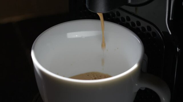 Espresso fills white cup from coffee machine, with warm brown stream and crema rising inside. Brewing process continues in close up, giving quiet cafe style mood and simple morning ritual.