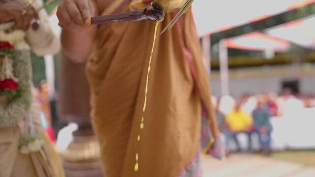 Hindu priest in traditional silk dhoti pours ghee into a sacred wedding fire while guests watch in the background, shown in slow motion.