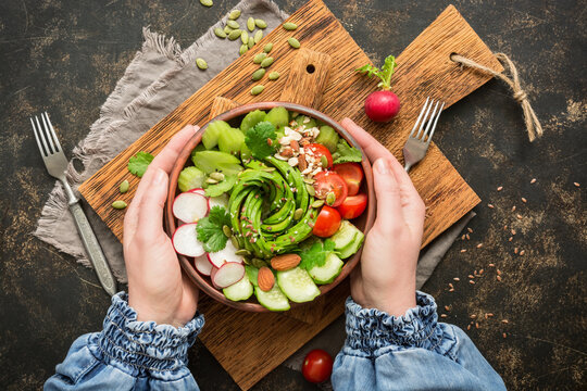 A bowl of fresh vegan vegetable salad held in a woman's hands on a rustic table, top view.