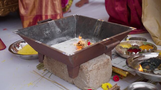 Close view of a priest's hand placing camphors into a metal fire pit during a Kannada wedding havan ritual with flowers and pooja items around.