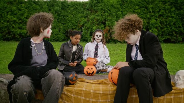 Diverse group of multiethnic teenagers in Halloween costumes sitting on lawn sharing candy from pumpkin buckets. Footage ideal for autumn holiday campaigns, confectionery ads, youth party promos