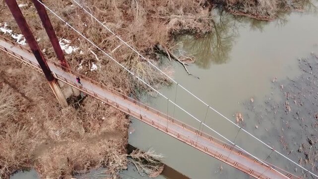 An old, rusty bridge over a murky river against an autumn backdrop, with a person walking across it.