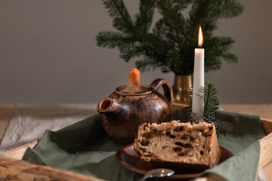 Piece of panettone cake on brown clay plate, crafted teapot, candle and pine tree branches on wooden tray, aesthetic cottage Christmas still life