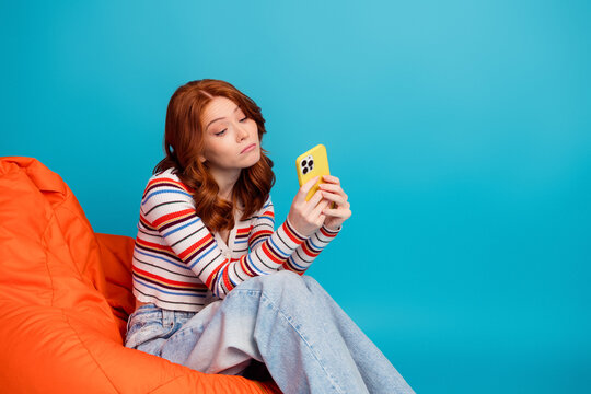 Young woman with red hair looking unimpressed at her smartphone while sitting on an orange bean bag against a bright blue background