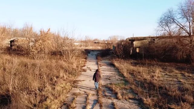 A person walking along an abandoned road made of reinforced concrete slabs, passing through the derelict buildings of an old factory.