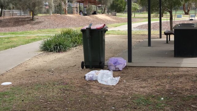 Overflowing garbage bin and littered plastic bags on bare dirt ground beside a park picnic shelter in Melbourne Australia. Littering, park maintenance neglect or public space cleanliness issues 