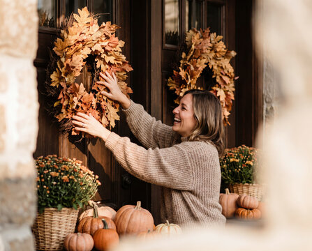 Playful woman hanging autumn leaf wreath among pumpkins on sunlit porch