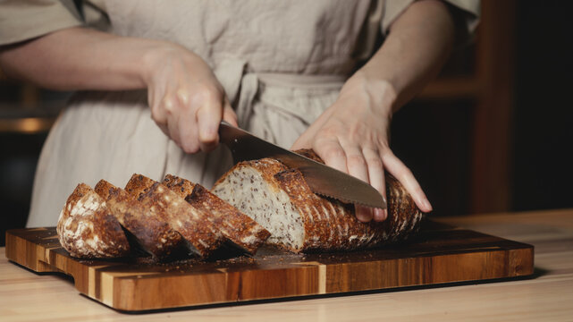 Close-up of baker in rustic linen apron slicing freshly baked artisan sourdough bread with knife on wooden cutting board. Homemade crusty loaf for breakfast, bakery promos, and healthy food concepts.