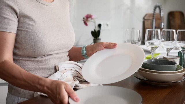 Woman drying plates and bowls with a cloth at a wooden kitchen table.