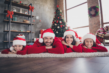 Naklejka premium Happy family lying on rug in matching Santa hats celebrating Christmas together at home with decorated tree and wrapped gifts