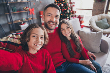 Naklejka premium Happy father and children wearing red sweaters taking a Christmas family selfie on a cozy living room couch with tree and gifts