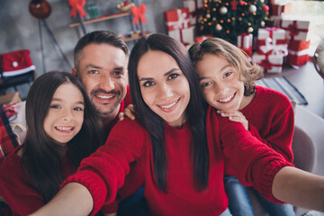 Naklejka premium Happy family of four wearing matching red sweaters celebrating Christmas together at home with gifts and decorated tree, parents and children smiling for a cozy holiday selfie