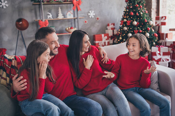 Naklejka premium Family wearing matching red sweaters enjoying Christmas together on a sofa by a decorated tree with wrapped gifts, sharing laughter and holiday warmth in a cozy living room