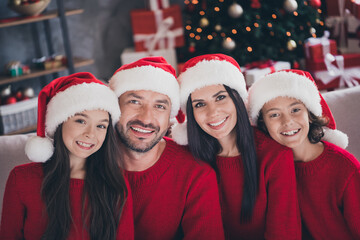 Naklejka premium Happy family wearing Santa hats smiling together on couch by decorated Christmas tree and wrapped gifts at home