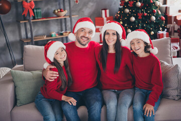 Naklejka premium Happy family wearing Santa hats sitting on couch by decorated Christmas tree enjoying holiday together at home