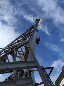 Low angle view of an industrial gas flare tower burning excess natural gas against a bright blue sky with wispy clouds.