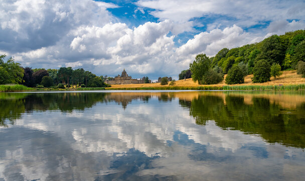 Panoramic view of Castle Howard across South Lake in North Yorkshire, England, surrounded by historic parkland and reflected in calm water beneath a dramatic summer sky.