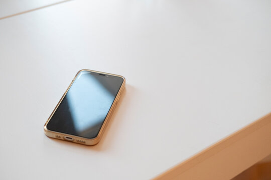 Smartphone resting on a clean white desk, modern minimal scene highlighting a sleek device, reflective screen, metal frame, soft ambient light, everyday technology at work.