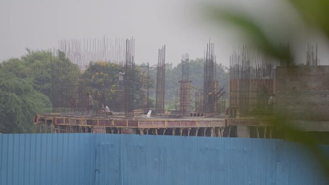 Concrete columns, reinforcing bars and wooden formwork rise behind a blue corrugated fence at an active construction site.