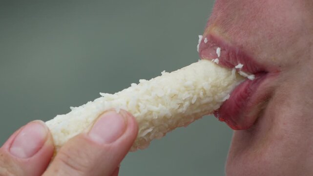 Macro view of person biting white coconut bar, chewing sweet snack outdoors. Static close-up shot.