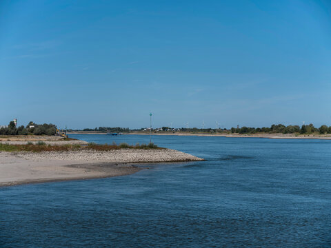 A wide Dutch river curves past a stone groyne and a pale sandbank, wind turbines standing on the far horizon.