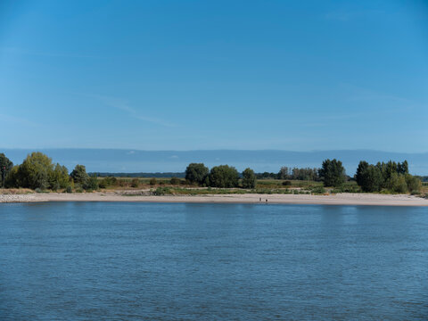 Low summer water leaves a broad sand beach along a Dutch river, a tree line and distant buildings beyond.