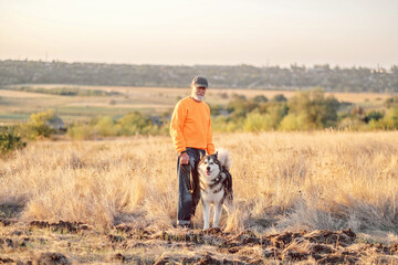 Man stands with large dog on open hill above quiet countryside village, countryside evening turning grass into gold, emotion easy to recognise. Wide shot. Concept of dog walking in autumn countryside © iwavephoto