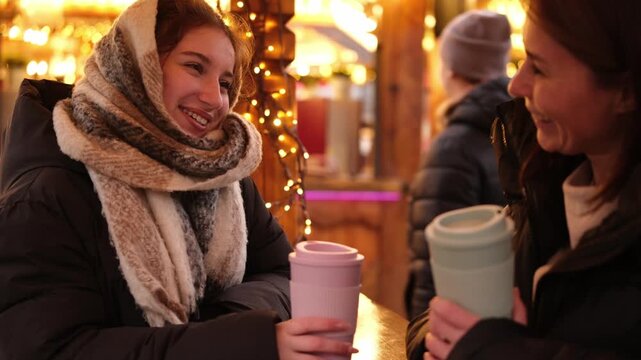 Girls Joking, Laughing, And Drinking Hot Holiday Wine At A Bright Christmas Fair