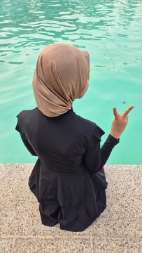 A woman wearing a beige hijab and black modest outfit sits beside a turquoise swimming pool and makes a peace gesture with her hand.