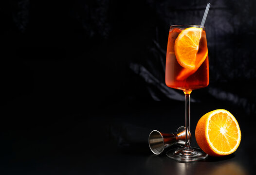 Aperol Spritz Cocktail with Orange. Classic Aperol Spritz cocktail in wine glass with ice cubes and orange slice, served with halved orange and bartending jigger on dark background.