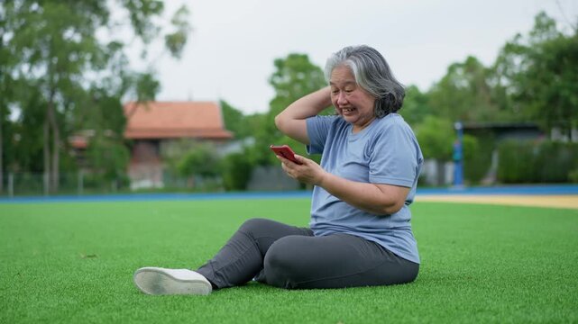 Older woman celebrates while using a smartphone outdoors on a sports field, raising her fist with a joyful expression for positive aging, digital connection, achievement and lifestyle storytelling.