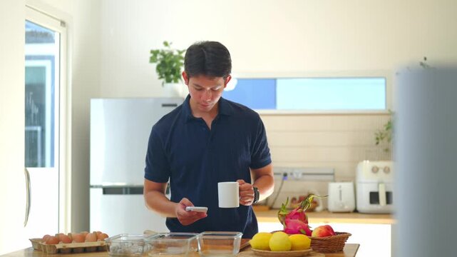 Young Asian Man Checking Smartphone While Holding Coffee Mug in Modern Kitchen Morning Routine